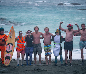 A group of eight expeditioners pose for the camera in front of the surf on Macquarie Island. One person has a surfboard, another is wearing a penguin suit and two men are flexing their arms.