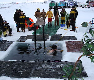 A man swims in a square hole cut in the ice as others wrapped up in warm clothing look on.