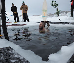 The head of a man just above water, swimming in an ice hole as others look on. A surf board and fake tree are in the background.