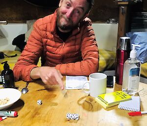 A man sitting at a table, head cocked to the size smiling to camera, pointing at three dice on the table