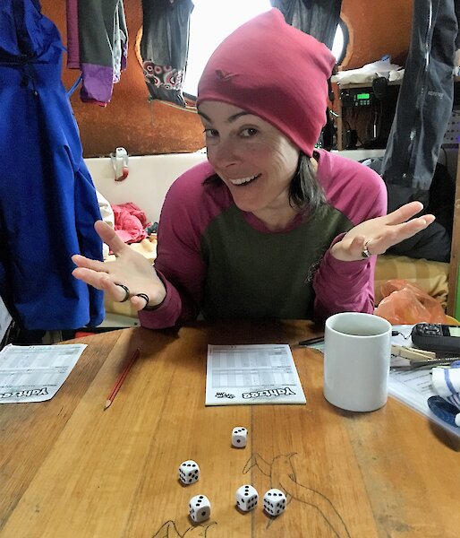 A women shrugs and smiles at camera, with five dice all showing three on the table in front of her