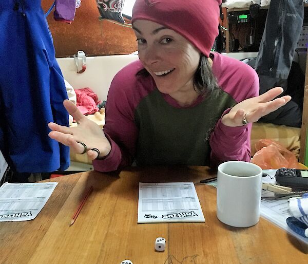 A women shrugs and smiles at camera, with five dice all showing three on the table in front of her