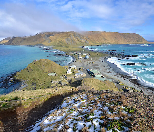 An aeriel view looking down a grassy hill, across a small isthmus with buildings on it, towards a grassy plateau