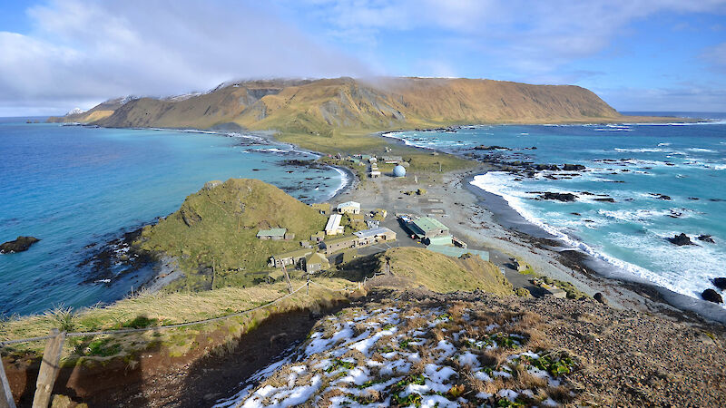 An aeriel view looking down a grassy hill, across a small isthmus with buildings on it, towards a grassy plateau