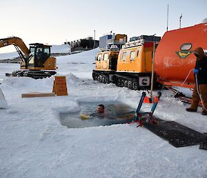 An expeditioner swimming in the finished ice hole with the yellow Hagglund and red caravan parked next to the ice hole.