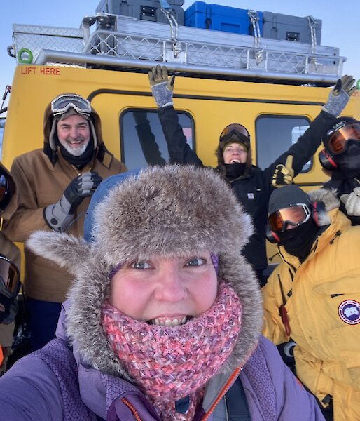 Selfie photo of excited expeditioners standing in front of the yellow Hagglund vehicle.