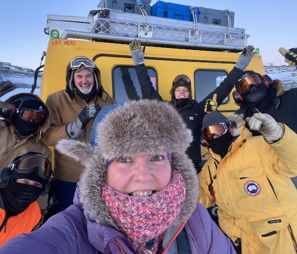 Selfie photo of excited expeditioners standing in front of the yellow Hagglund vehicle.