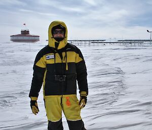 Man in yellow and black windproof gear stands on snow with station building and wind turbines in background.