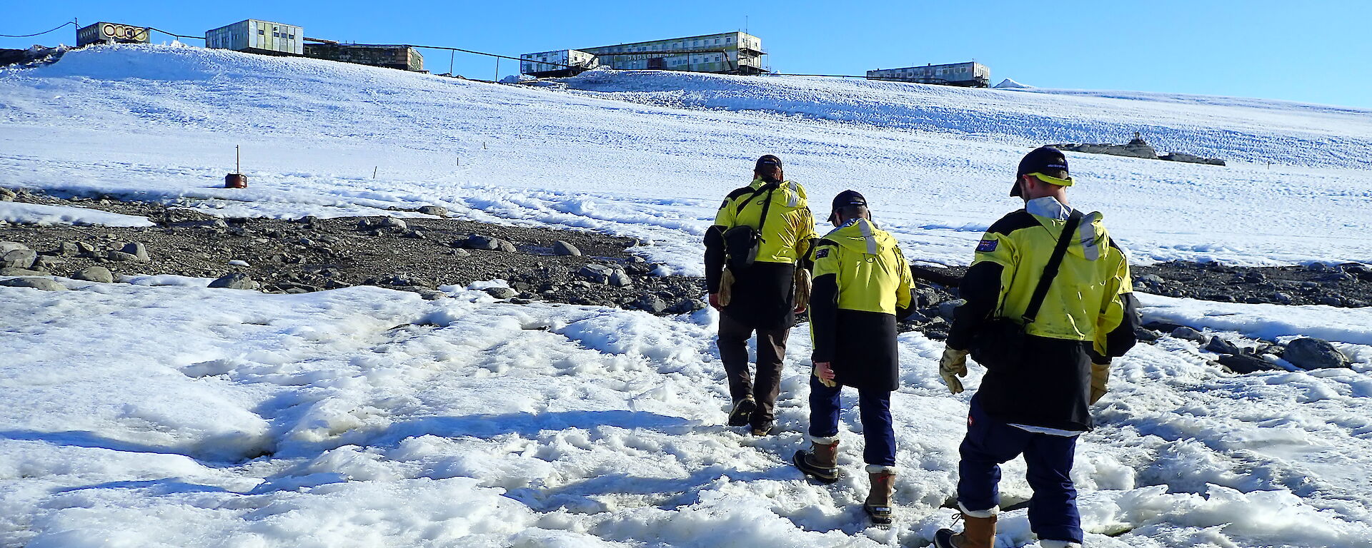 Three people in yellow jackets waslking across snow to distant box-shaped buildings.