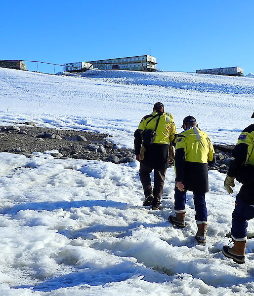 Three people in yellow jackets waslking across snow to distant box-shaped buildings.