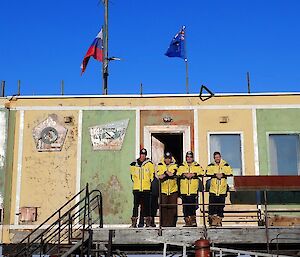 Four men outside aged square building with flags on roof.