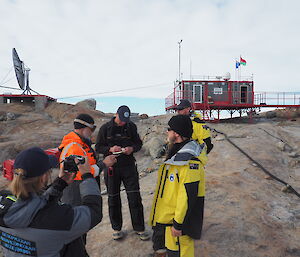 Group of men talking on rocky ground with buildings in background.