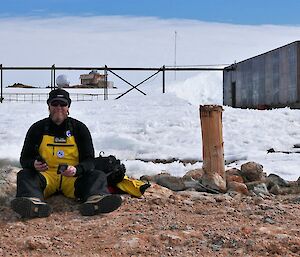 Man sits of rocky ground with buildings and snow in background.