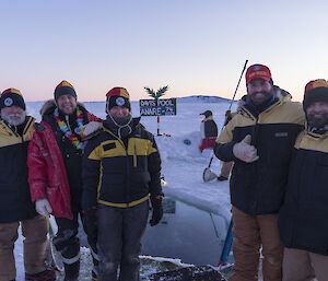 A group stand and smile to camera in front of an ice-hole.  A 'Davis Pool' sign is erected in the background.