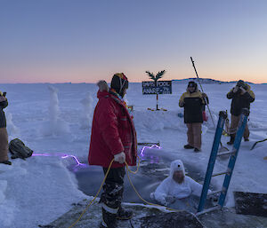 A man in a polar bear suit submerged in an ice hole with a crowd around him.  A sign at the back reads Davis Pool ANARE '74 2021