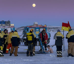 A man enters an ice pool pool with a crowd, in warm weather gear, watching him.  A full moon sits high above in a pink sky