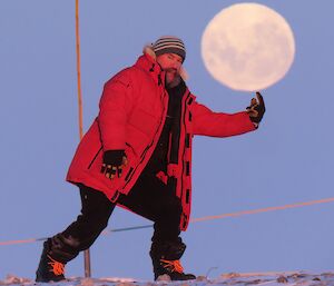 A man standing with his finger pointed upwards to look as if he is balancing the moon, in the sky behind him, on his finger