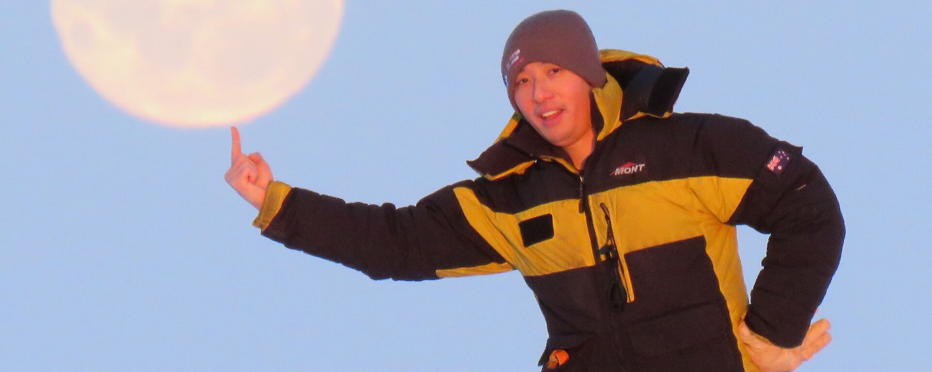 A man standing with his finger pointed upwards to look as if he is balancing the moon, in the sky behind him, on his finger