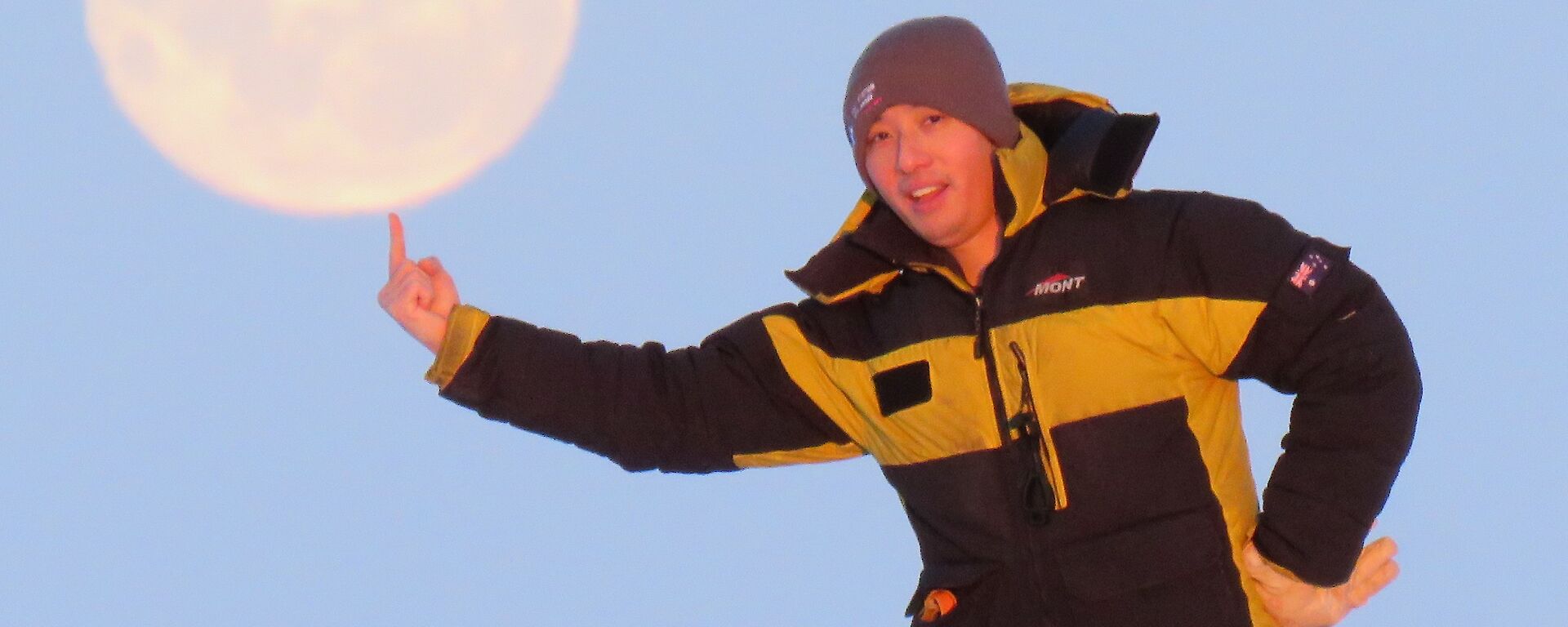 A man standing with his finger pointed upwards to look as if he is balancing the moon, in the sky behind him, on his finger