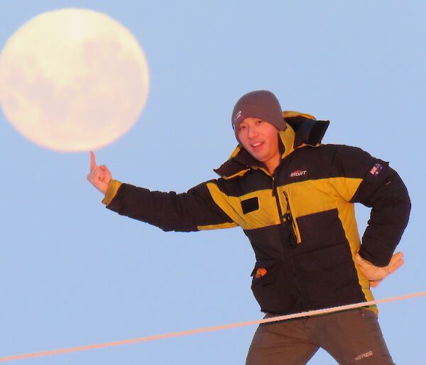 A man standing with his finger pointed upwards to look as if he is balancing the moon, in the sky behind him, on his finger
