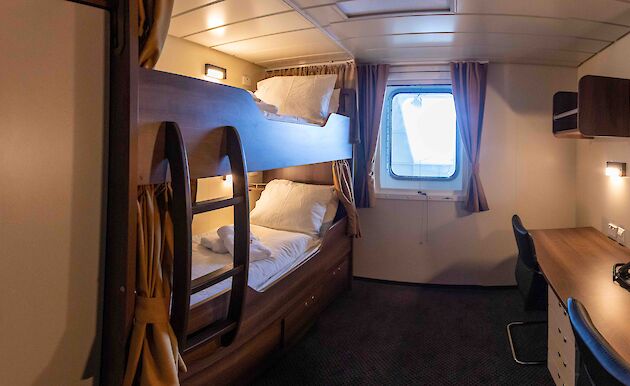 One of the ship's cabins with a bunk bed and desk either side of a port hole.