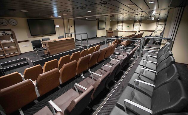 Rows of padded seats looking towards a large screen TV and desk on a raised level.