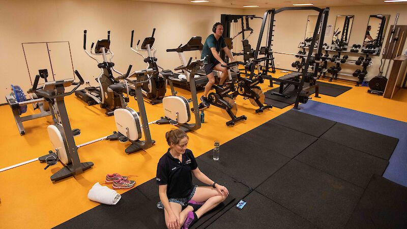 A woman sitting on a yoga mat and a man pedalling an exercise bike, surrounded by other bikes and gym equipment.