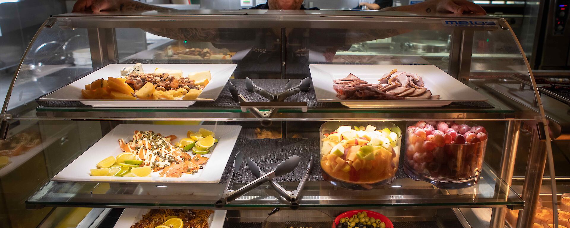 Galley staff show off platters of fruit, meat, cheeses and salads on serving plates.
