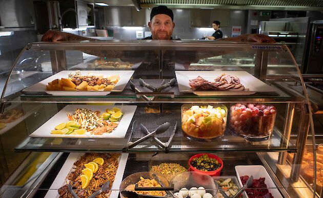 Galley staff show off platters of fruit, meat, cheeses and salads on serving plates.