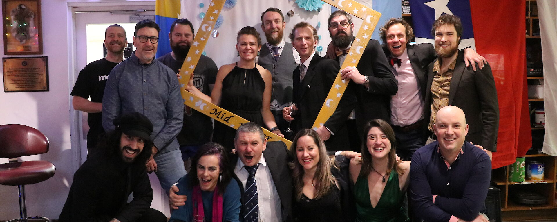 A group shot of people dressed up for a party, some holding an oversized picture frame, all smiling to camera