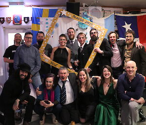 A group shot of people dressed up for a party, some holding an oversized picture frame, all smiling to camera