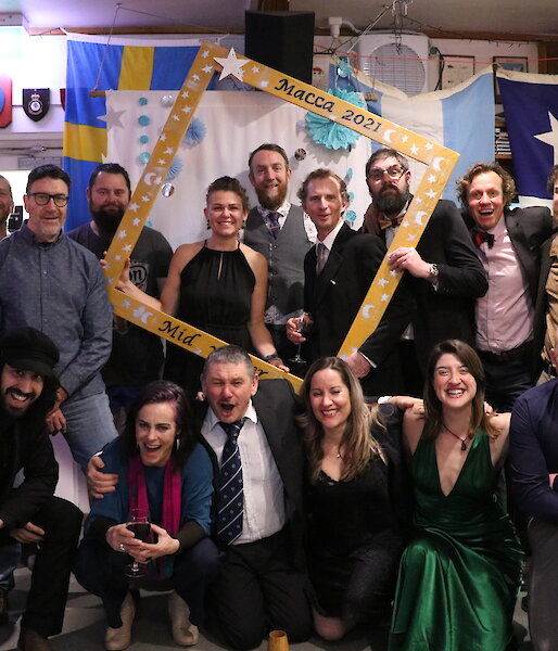 A group shot of people dressed up for a party, some holding an oversized picture frame, all smiling to camera