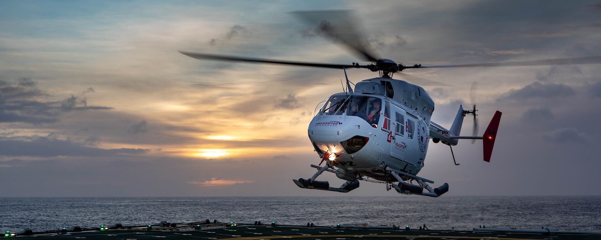 A white and red helicopter lands on a ship's helicopter deck at dusk