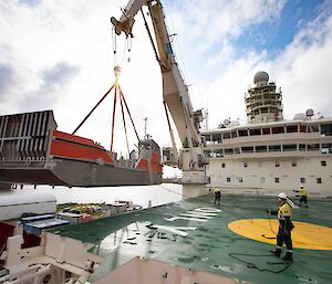 Orange and silver barge being lifted by crane onto deck of ship. Two workers are holding ropes to guide it into place.