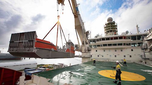 Orange and silver barge being lifted by crane onto deck of ship. Two workers are holding ropes to guide it into place.