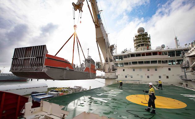 Orange and silver barge being lifted by crane onto deck of ship. Two workers are holding ropes to guide it into place.