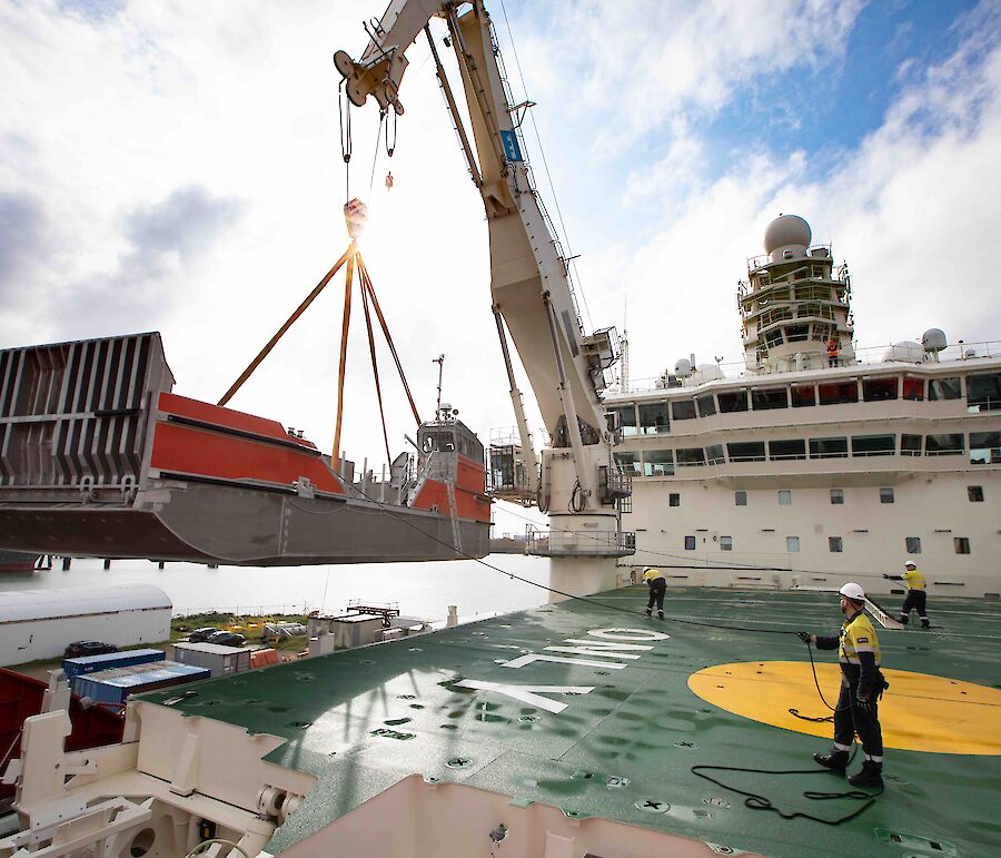 Orange and silver barge being lifted by crane onto deck of ship. Two workers are holding ropes to guide it into place.