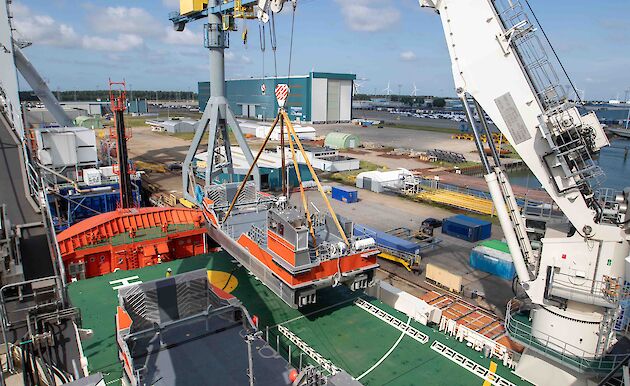 View from above of orange and silver barge being lifted by crane onto deck of ship to rest beside an identical barge.