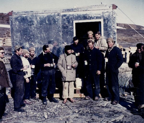 A group of expeditioners standing in front of a shiny new building having a hot drink.