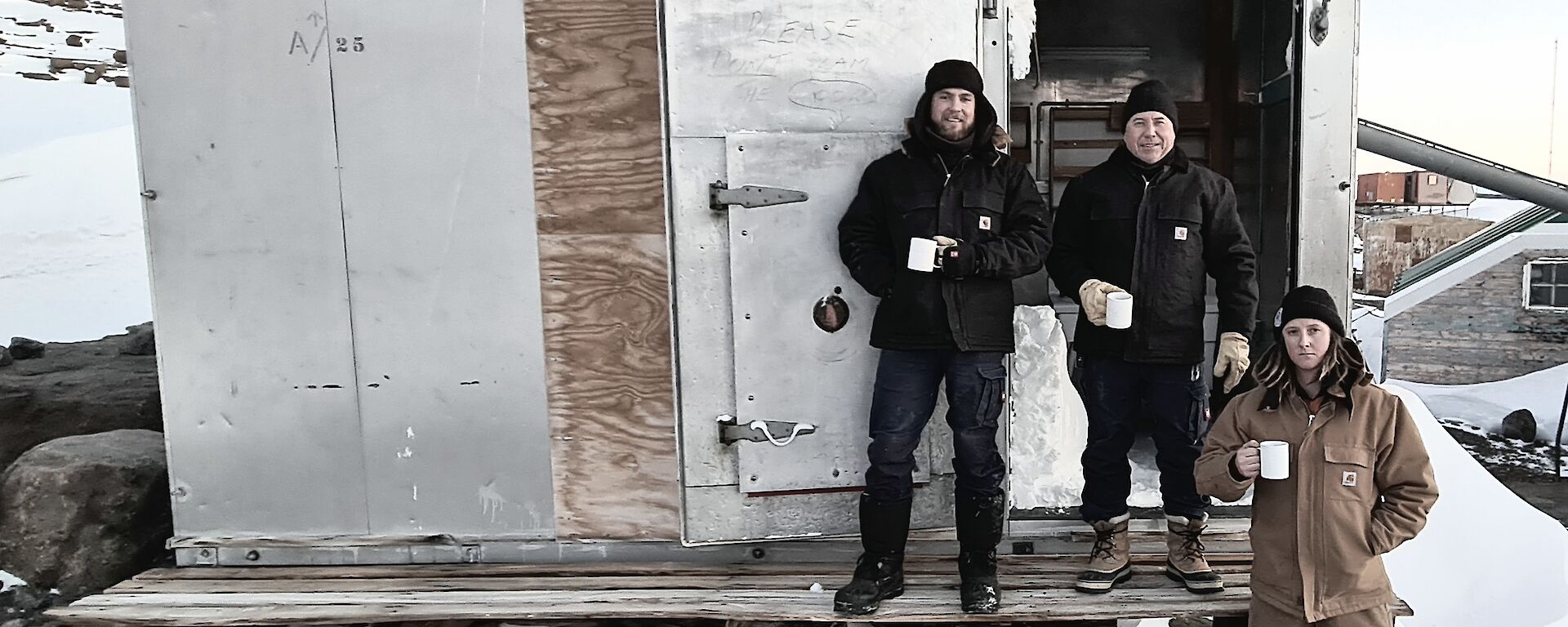 A metal and hardwood hut with three men standing out front having a cup of tea