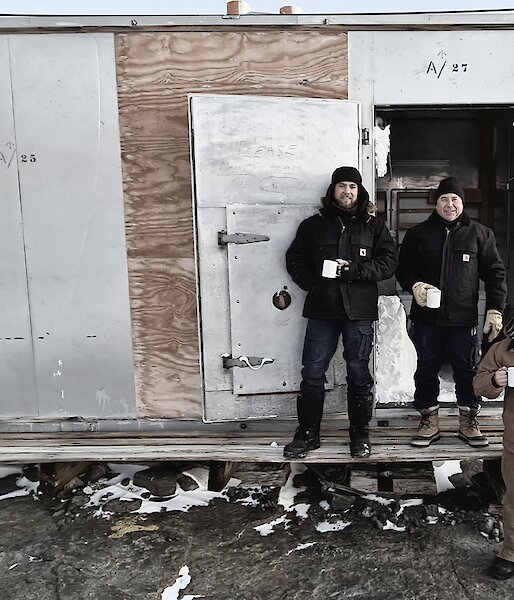 A metal and hardwood hut with three men standing out front having a cup of tea