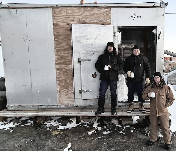 A metal and hardwood hut with three men standing out front having a cup of tea
