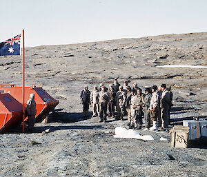 A group of men standing in front of the Australian flag on the rocky location that Mawson station would be built.