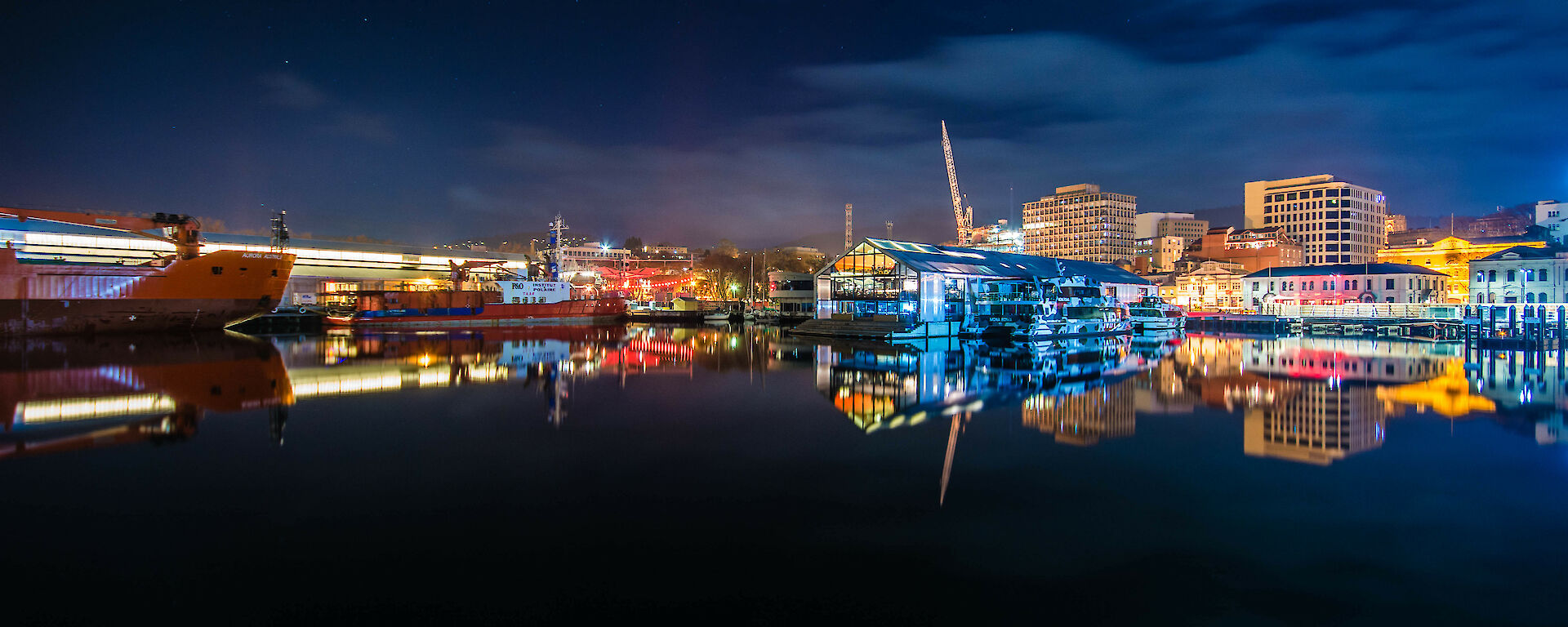 Night-time shot of ships and colourfully lit buildings reflecting on still, black water.