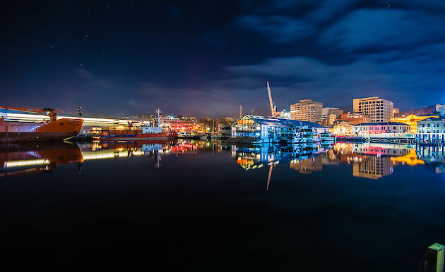 Night-time shot of ships and colourfully lit buildings reflecting on still, black water.