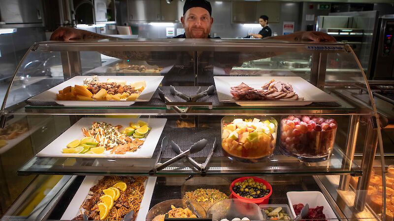 Galley staff show off a delicious spread of food including salads, meats, cheeses and fruit and serving bowls.