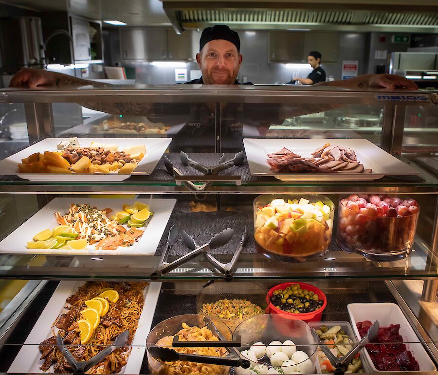 Galley staff show off a delicious spread of food including salads, meats, cheeses and fruit and serving bowls.