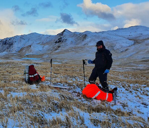 A man on Macquarie Island, standing beside two back packs and a solar panel, with snow covered grass and mountains in the distance.