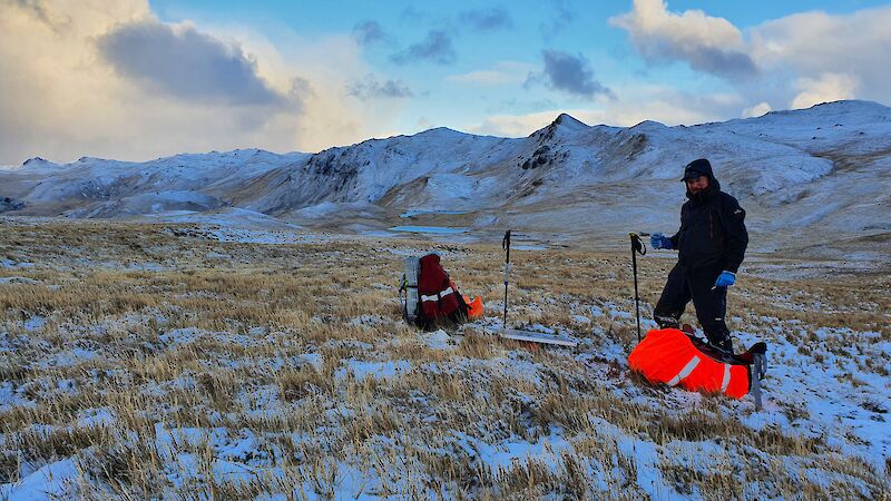 A man on Macquarie Island, standing beside two back packs and a solar panel, with snow covered grass and mountains in the distance.