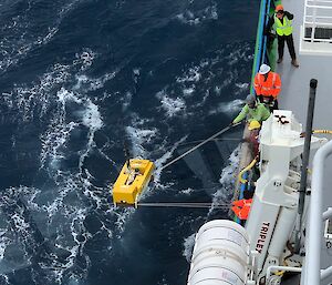 A scientific instrument being craned over the side of a ship into the ocean with 5 people watching on.